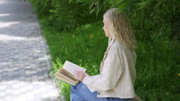 A Young Blonde Woman with Curls Sits on a Park Bench and Reads a Fascinating Book