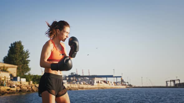 Young Girl in Boxing Gloves and Sport Clothes is Boxing While Training By Seaside Near the City Port alt