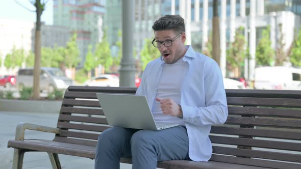 Man Celebrating Success on Laptop While Sitting Outdoor on Bench alt