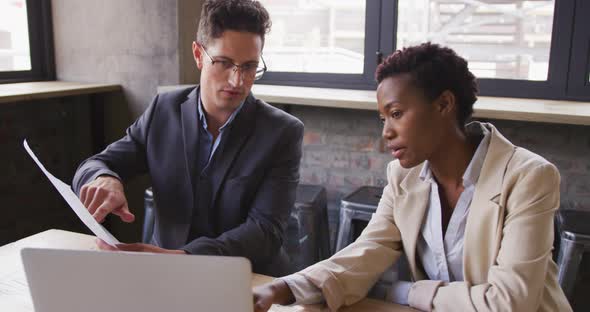 Diverse male and female business colleagues sitting talking in cafe looking at laptop and paperwork alt