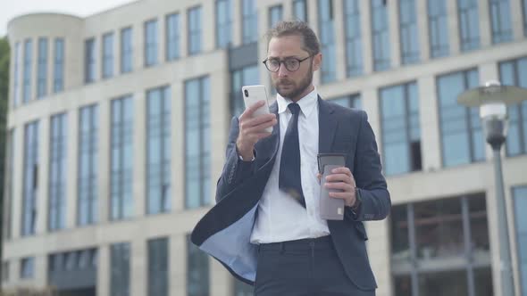 Portrait of Concentrated Nerd Businessman Using Smartphone Outdoors in Front of Office Business alt