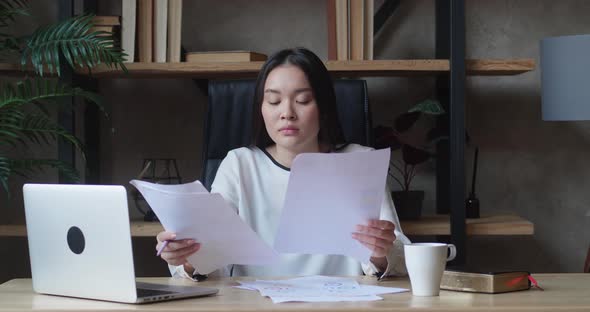 Stressed Unmotivated Asian Woman Confused By Mistake in Documents Looking Through Papers Frowning alt