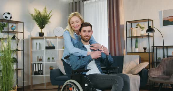 Disabled Man in Wheelchair which Looking at Camera with His Caring Smiling Light-Haired Wife alt