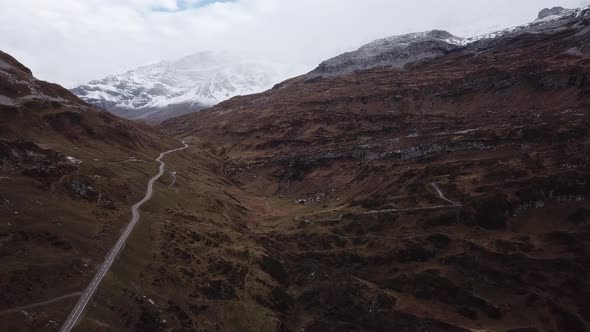 Aerial shot of a rocky mountain landscape criss-crossed by winding roads and snowy hills in the back alt
