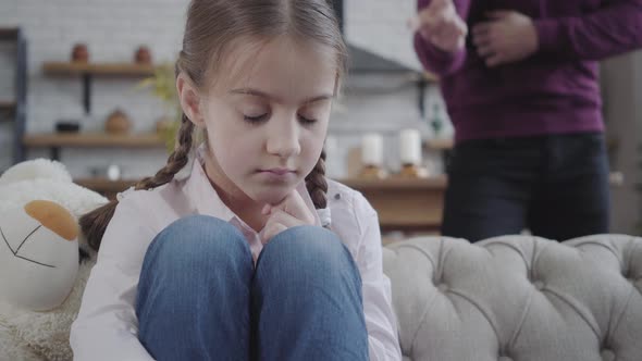 Close-up of Cute Caucasian Brunette Girl with Braided Pigtails Sitting on Sofa and Hugging Knees alt