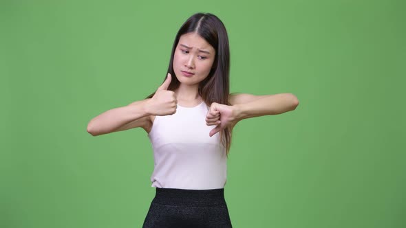 Young Confused Asian Businesswoman Choosing Between Thumbs Up and Thumbs Down