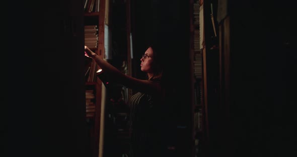 Girl in Glasses with a Lantern Looks for a Book on Library Shelves in Darkness alt
