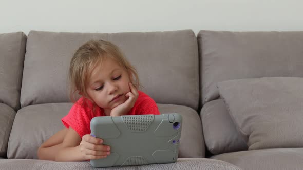 Portrait of Excited Little Girl Using Digital Tablet While Lying on Sofa Watching Cartoons or