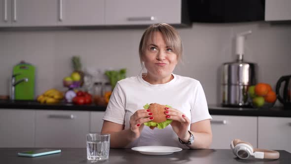 Hungry Woman Eating Burger Sitting in Kitchen alt