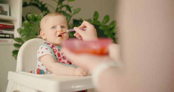 Little baby girl eating baby food on high chair with mother alt