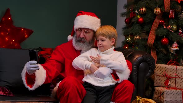 Santa Claus with a Child Sitting on His Lap are Taking a Selfie Using a Smartphone alt