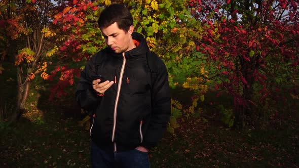 Young Man in Black Jacket in the Autumn Park Using His Smartphone alt