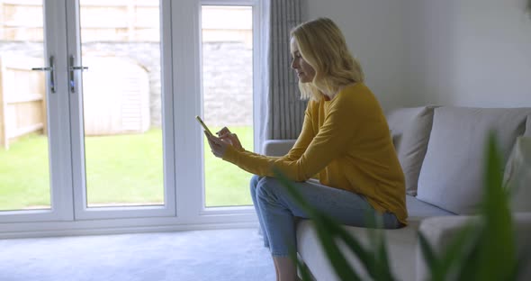 Blond woman sitting on couch scrolling on smartphone