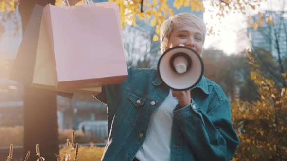 Young Blond Woman with Shopping Bags Announcing Sales Season with Loudpeaker in the City Park in alt