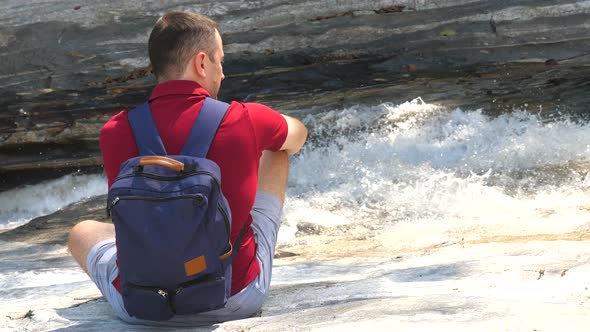 Alone Male Traveler with a Backpack Sits Near a Bubbling Mountain River alt