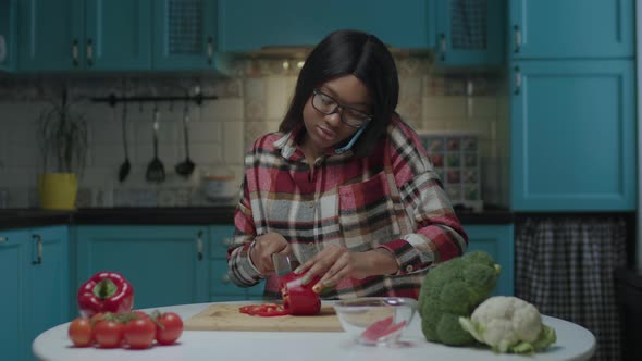 20s African American Woman Talking By Mobile Phone and Cooking Fresh Vegetable Salad Sitting at the alt