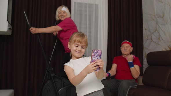 Senior Couple Using Orbitrek Doing Dumbbells Exercises Child Girl Making Selfie on Mobile Phone alt