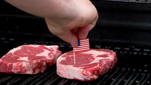 Two juicy rib-eye steaks sitting on the grill and cooking a hand comes in and sticks a tiny American alt