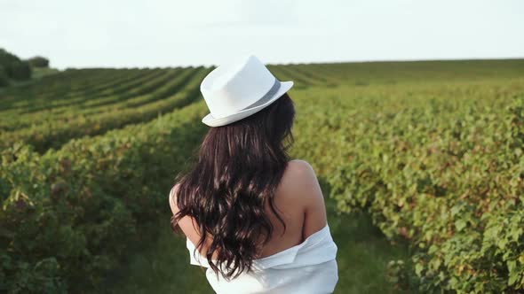 Beautiful Girl in Hat Walks Among Bush Plantation and Turns to Camera with Smile alt