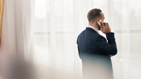 Male Businessman Answer for Incoming Call Using Touchscreen of Smartphone Standing in Front Window alt