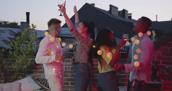 Company Dancing with Glasses of Wine and Yellow Lighted Bulbs on Summer Party on Terrace alt