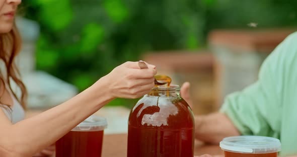 Tasting of Fresh Honey in the Apiary. Beekeeper Opens the Lid on a Jar of Honey, the Girl Tries the alt