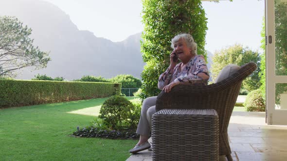 Caucasian senior woman smiling while talking on smartphone while sitting on the chair outdoors alt