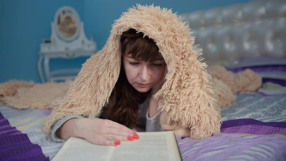 Brunette Woman Lies on the Bed and Reads a Book Tited and Covered with a Blanket