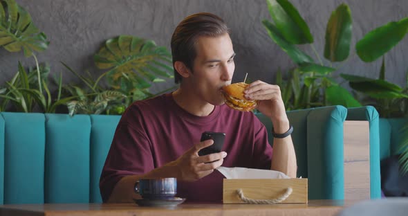 Man Eating Burger and Sliding Smartphone While Having a Coffee Break in Small Cafe alt