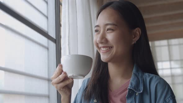 Smiling young Asian woman holding coffee cup standing beside a window at the home office.