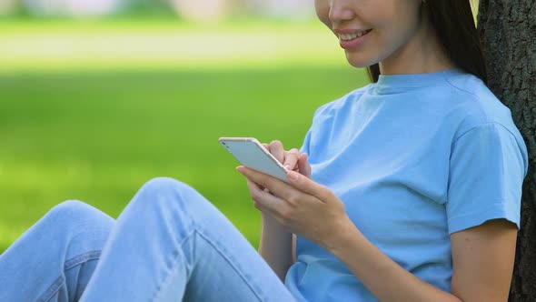 Young Smiling Female Chatting on Smartphone, Relaxing in Sunny Day in Park alt