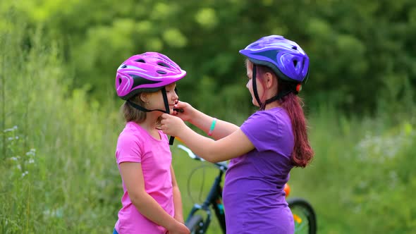 The older sister puts on the younger one a protective bicycle helmet in the park in the summer