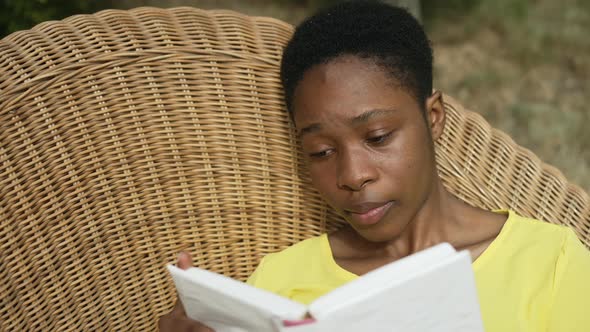 Closeup Absorbed African American Beautiful Woman Reading Book Lying on Wicker Sofa in Garden alt
