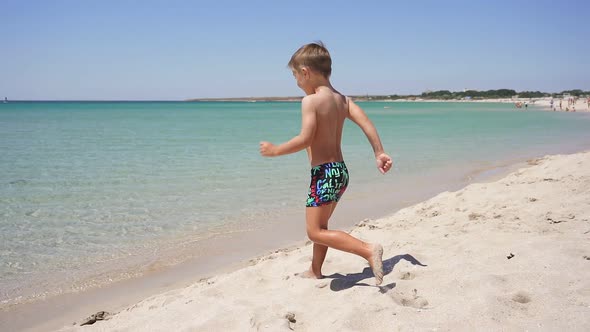 Little Boy Runs Along a Sandy Beach Directly Into the Sea on a Sunny Day Raising Spray
