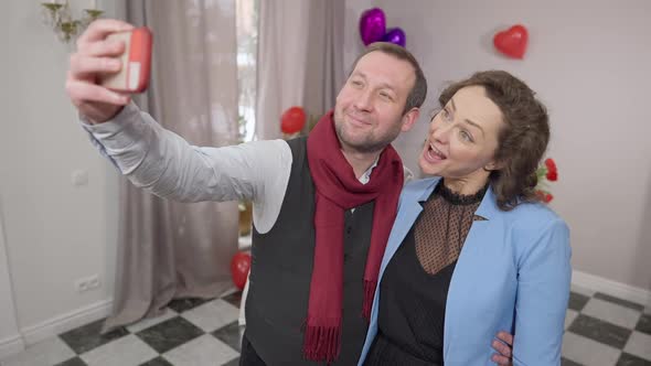 Positive Funny Adult Couple Making Faces Taking Selfie Indoors on Valentine's Day alt