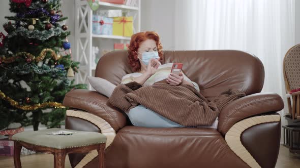 Ill Tired Caucasian Woman in Coronavirus Face Mask Using Smartphone and Sneezing Sitting at Home alt