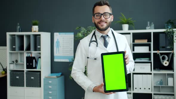 Cheerful Young Man Doctor in White Gown Is Holding Tablet with Green Chroma Key Screen Smiling alt