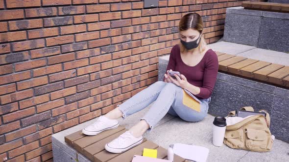 Female with Protective Mask Surfs Internet with Phone on Bench