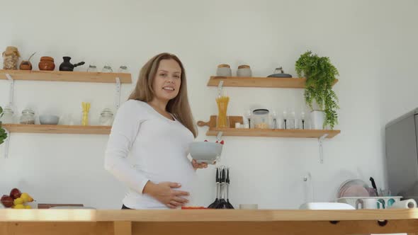 Pregnant Fairskinned Woman in the Kitchen Holding a Bowl of Chopped Red Peppers alt