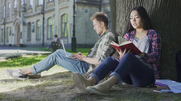 Multiracial Girl Closing Book, Pressing It to Chest, Looking at Guy With Laptop alt