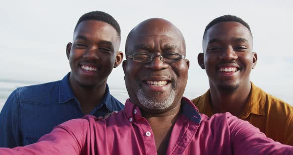 Smiling african american senior father and twin teenage sons standing on a beach taking a selfie alt