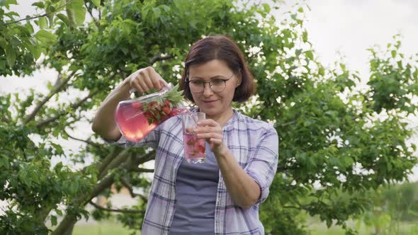 Middle Aged Woman Pouring Natural Drink with Strawberries and Mint From Jug Into Glass alt