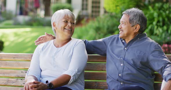 Video of happy biracial senior couple embracing and sitting on bench in garden alt