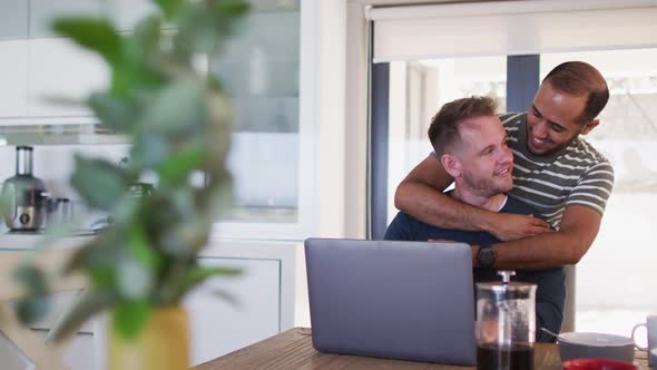 Multi ethnic gay male couple embracing in kitchen one sitting using laptop alt