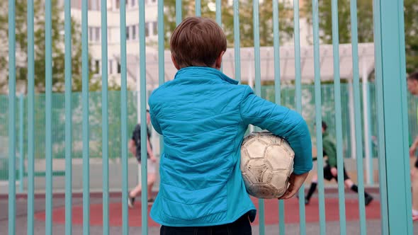 A Little Insecure Boy Holding Deflated Ball and Watching Other Kids Playing Football alt