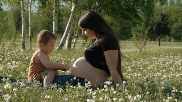 A Young Pregnant Woman and Her Son Are Sitting Happily on a Flower Meadow alt