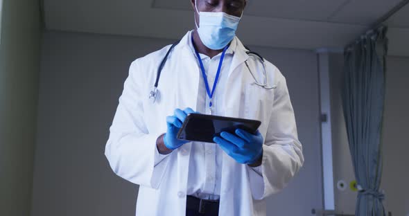 African american male doctor wearing face mask using digital tablet in hospital patient room alt