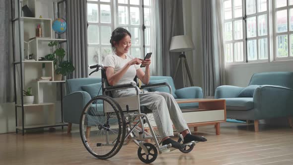 Asian Woman Sitting In A Wheelchair While Smiling And Using Smart Phone At Home alt