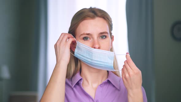Young woman putting on a medical blue mask at home alt