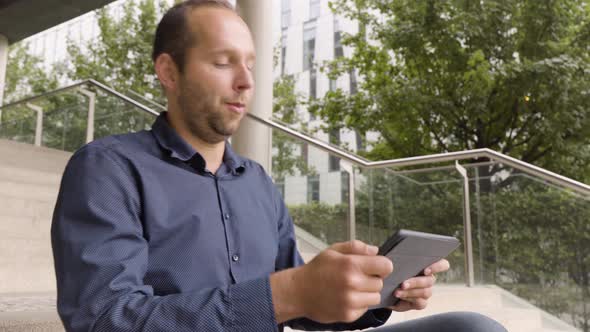 A Caucasian Man Plays a Game on a Tablet As He Sits on a Staircase in an Urban Area